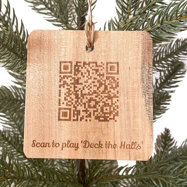 Hand made wood ornament with a QRCode laser engraved and the text 'Scan to play 'Deck the Halls'' on a pine tree background.