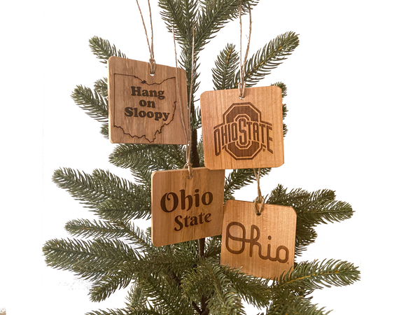 Four OSU Ohio State Ornaments - Fallen wood showing natural wood grain, laser engraved designs - 1 Hang on Sloopy text - to the right Ohio State sport logo - below - Script Ohio - below left Ohio State text - on pine tree background.