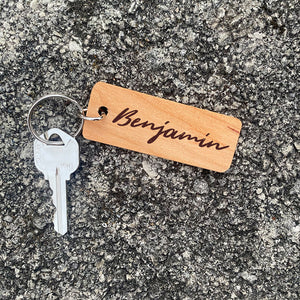 Handcut wood keychain with laser engraved name "Benjamin" with a silver key on a concrete background.