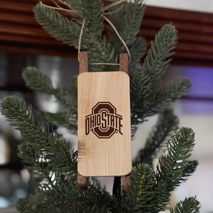 Light colored wood sled ornament with OSU sport logo laser engraved design, hung on an artificial green pine tree. 