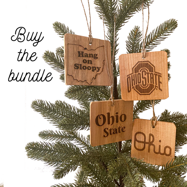 Four OSU Ohio State Ornaments - Fallen wood showing natural wood grain, laser engraved designs - 1 Hang on Sloopy text - to the right Ohio State sport logo - below - Script Ohio - below left Ohio State text - on pine tree background.