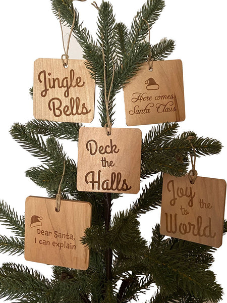 Five hand crafted wood ornaments hanging on a pine tree with laser engraved text. From left to right, top to bottom, Jingle Bells, Here comes satan Claus, Deck the Halls, Dear Santa I can Explain, Joy to the world.