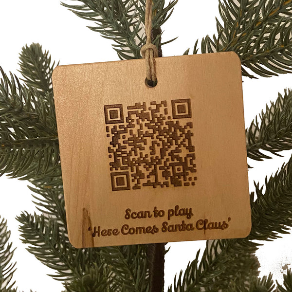 Hand crafted wood ornament with a laser engraved QRCode and text below "Scan to play 'Here Comes Santa'" on a pine tree background. 