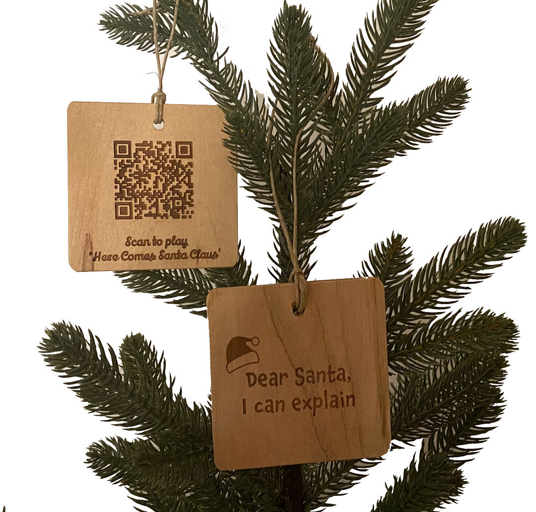 Two hand crafted wood ornaments with laser engraved designs handing on a small pine tree with a white background. Ornament on the left has a QR Code to be able to scan and play the song "Here comes Santa." The right ornament has a laser engraved of a Santa hat and the text "Dear Santa, I Can Explain."