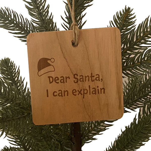 Hand made wood ornament with laser engraved of a Santa hat and the text "Dear Santa, I Can Explain." On a pine tree background.