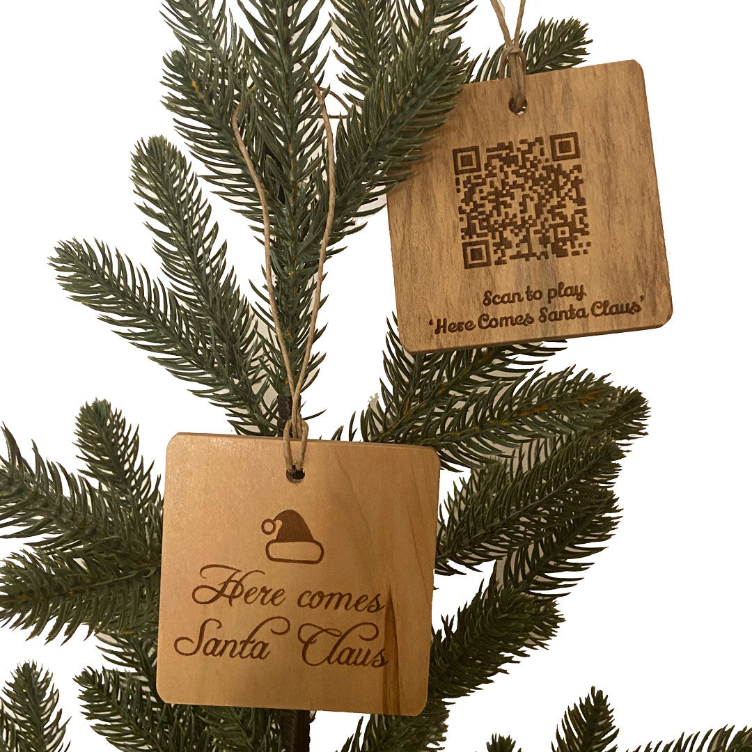 Two hand crafted wood ornaments with laser engraved designs handing on a small pine tree with a white background. Ornament on left has Here Comes Santa design and Ornament on the right has a QR Code to be able to scan and play the song.