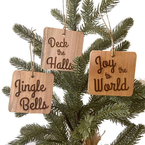 Three hand crafted wood ornaments hanging on a pine tree with laser engraved text. One with Jingle Bells text, the middle with Deck the Halls text and the right with Joy to the World Text