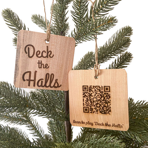 Two hand crafted wood ornaments with laser engraved designs handing on a small pine tree with a white background. Ornament on left has Deck the Halls design and Ornament on the right has a QR Code to be able to scan and play the song.