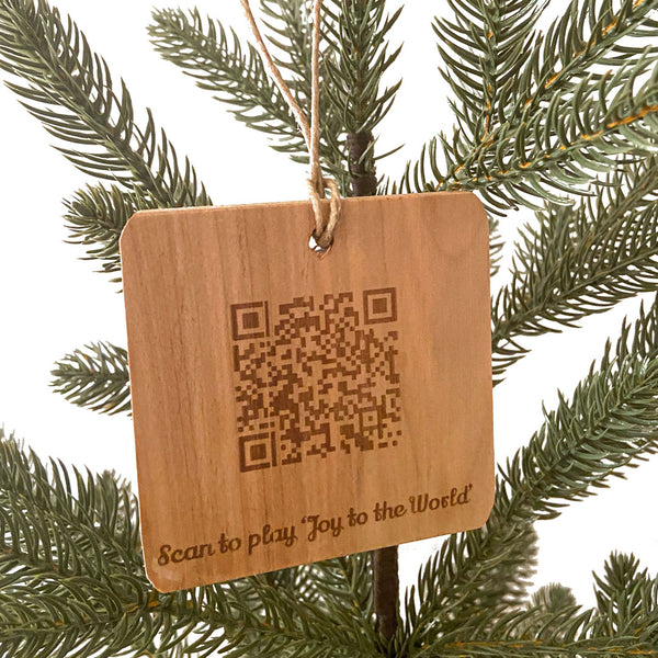 Hand crafted wood ornament hanging on a pine tree with a white background. Laser engraved design QRCode to scan and play the song 'Joy to the world.'