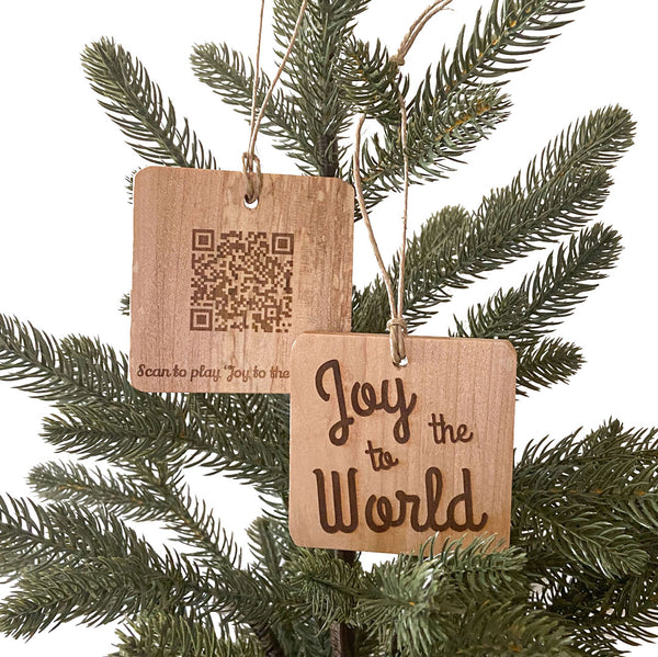 Two hand crafted wood ornaments with laser engraved designs handing on a small pine tree with a white background. Ornament on right has Deck the Halls design and Ornament on the left has a QR Code to be able to scan and play the song.