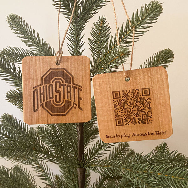 Two hand cut wood ornaments laser engraved OSU Sport logo design on one and and on the back a QR Code to play the song 'Across the Field.' Hanging from a pine tree with white background.