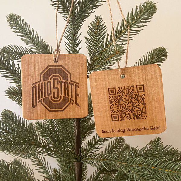 Two hand cut wood ornaments laser engraved OSU Sport logo design on one and and on the back a QR Code to play the song 'Across the Field.' Hanging from a pine tree with white background. 