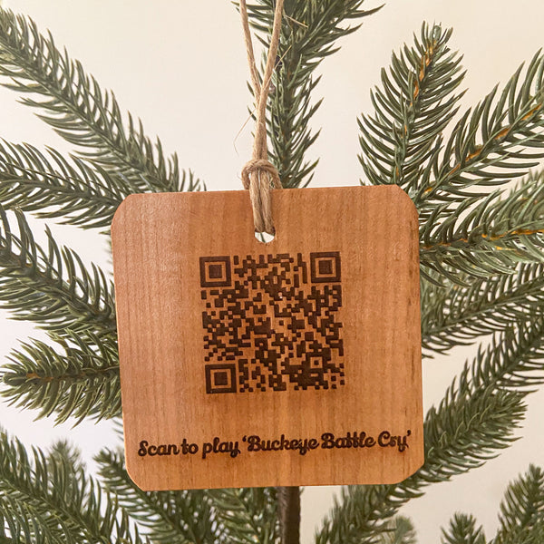 Natural wood ornament, laser engraved QR Code to scan and play 'Buckeye Battle Cry'. Hanging from a pine tree.