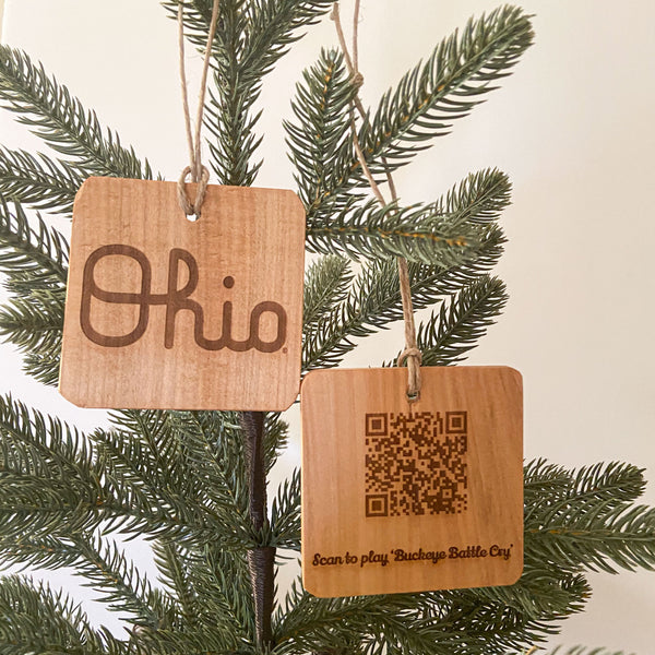 Natural wood ornament with laser engraved OSU Script Ohio. Next to it another ornament showing the back, laser engraved QR Code to scan and play 'Buckeye Battle Cry'. Hanging from a pine tree.