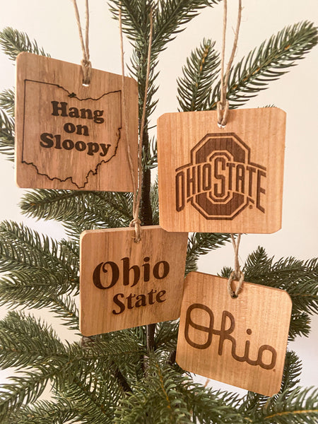 Four OSU Ohio State Ornaments - Fallen wood showing natural wood grain, laser engraved designs - 1 Hang on Sloopy text - to the right Ohio State sport logo - below - Script Ohio - below left Ohio State text - on pine tree background.