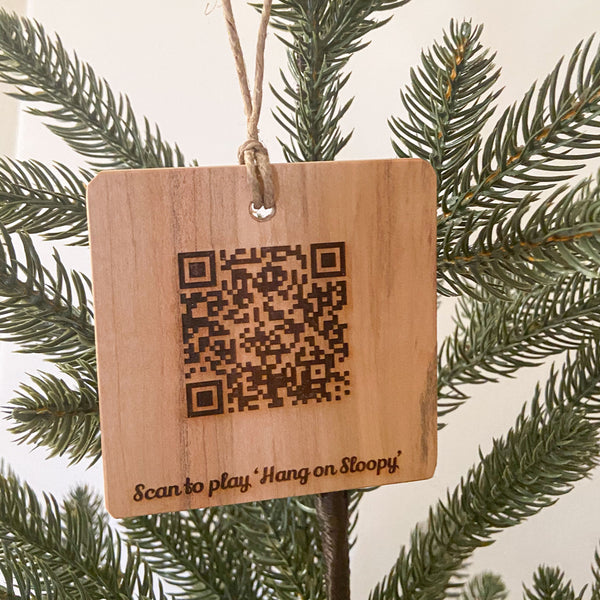 QR Code scannable natural wood ornament with text Scan to play 'Hang on Sloopy'. Hanging from a pine tree with a white background.