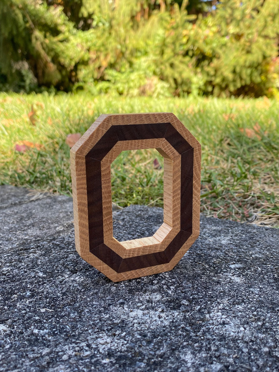 Freestanding handmade wood block O, with dark wood in the center. Set on a concrete background.