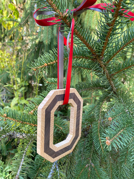 From an angle, hand cut original Block O Ornament tied with scarlet and gray ribbon hanging on a pine tree.
