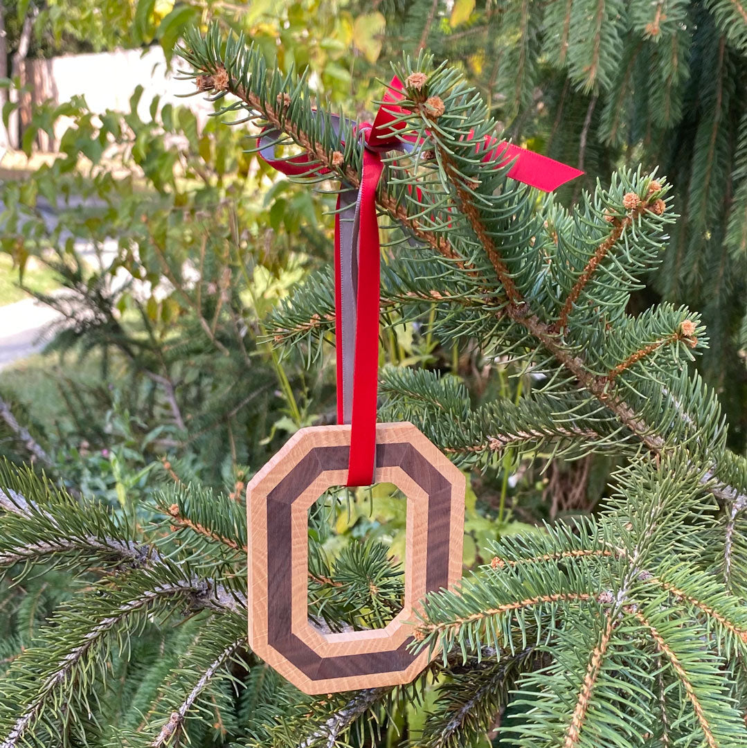 Hand cut original Block O Ornament tied with scarlet and gray ribbon hanging on a pine tree.