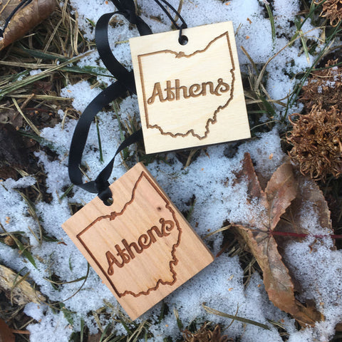 Wood ornament with laser engraved design, Athens text enclosed in the shape of Ohio. Snow and leaves in background.