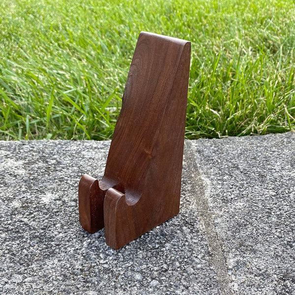 Walnut wood phone stand with charging access on concrete with grass background.