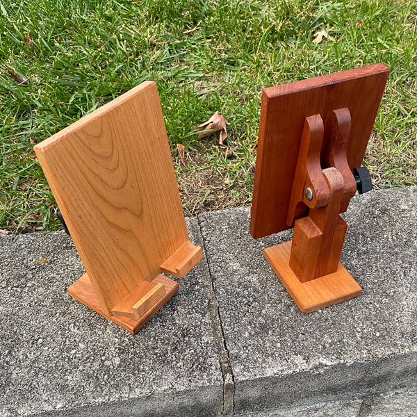 Two adjustable wood tablet stand. One facing the front and one facing the back, looking down on them. A grass and concrete background.