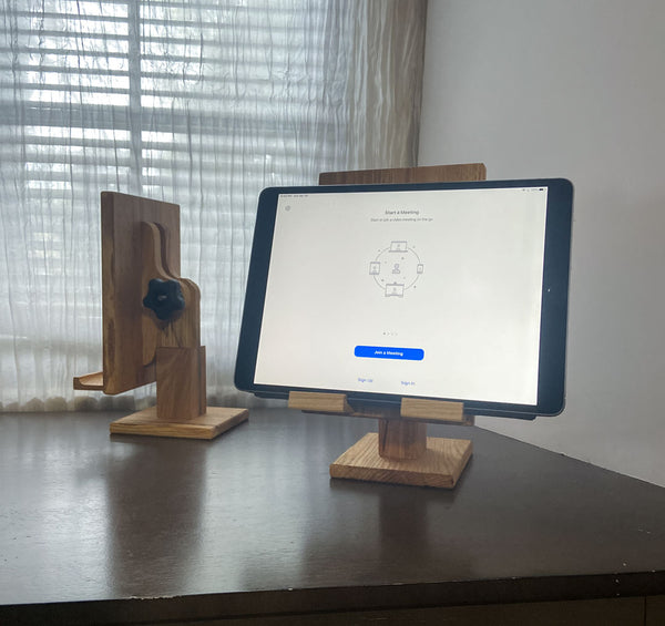 Two adjustale wood tablet stands, one facing front with an iPad displaying a zoom call. The one in the back shows the adjustable knob. White curtain background.