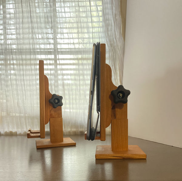 Two adjustable wood tablet stands at a strait angle. One with an ipad on it and one without. A sheer curtained window in the background.