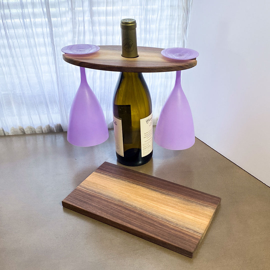 Wood walnut wine and glass display shown with two purple wine glasses hanging. Set in a wine bottle. With a coordinating cheese board in front.