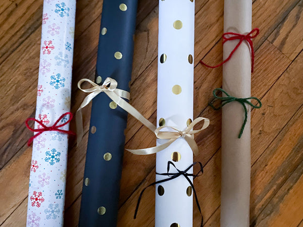 4 rolls of wrapping paper laying on a wood hardwood floor tied with ribbon. REd, Green and Pink snowflake paper, black with gold dots, white with golds dots and craft brown paper.