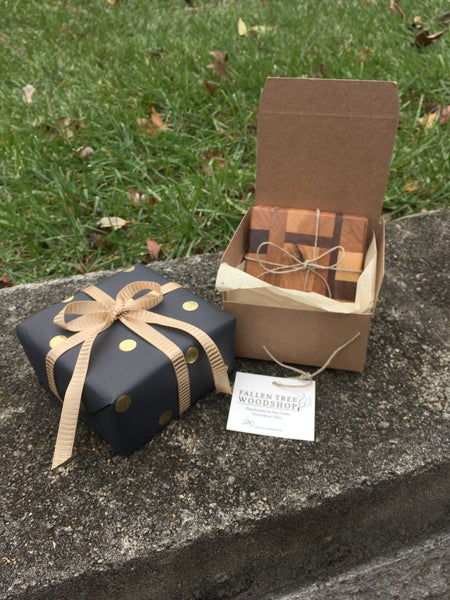A small square box wrapped in black paper with gold dots tied in a gold ribbon. Next to it a brown box open with a handmade wood coaster set tied in twine.