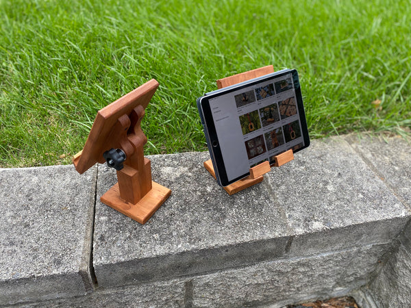 Two adjustable tablet stands shown at an angle, one facing the front and one facing in the back. The one in the front has an ipad on it.Grass background.